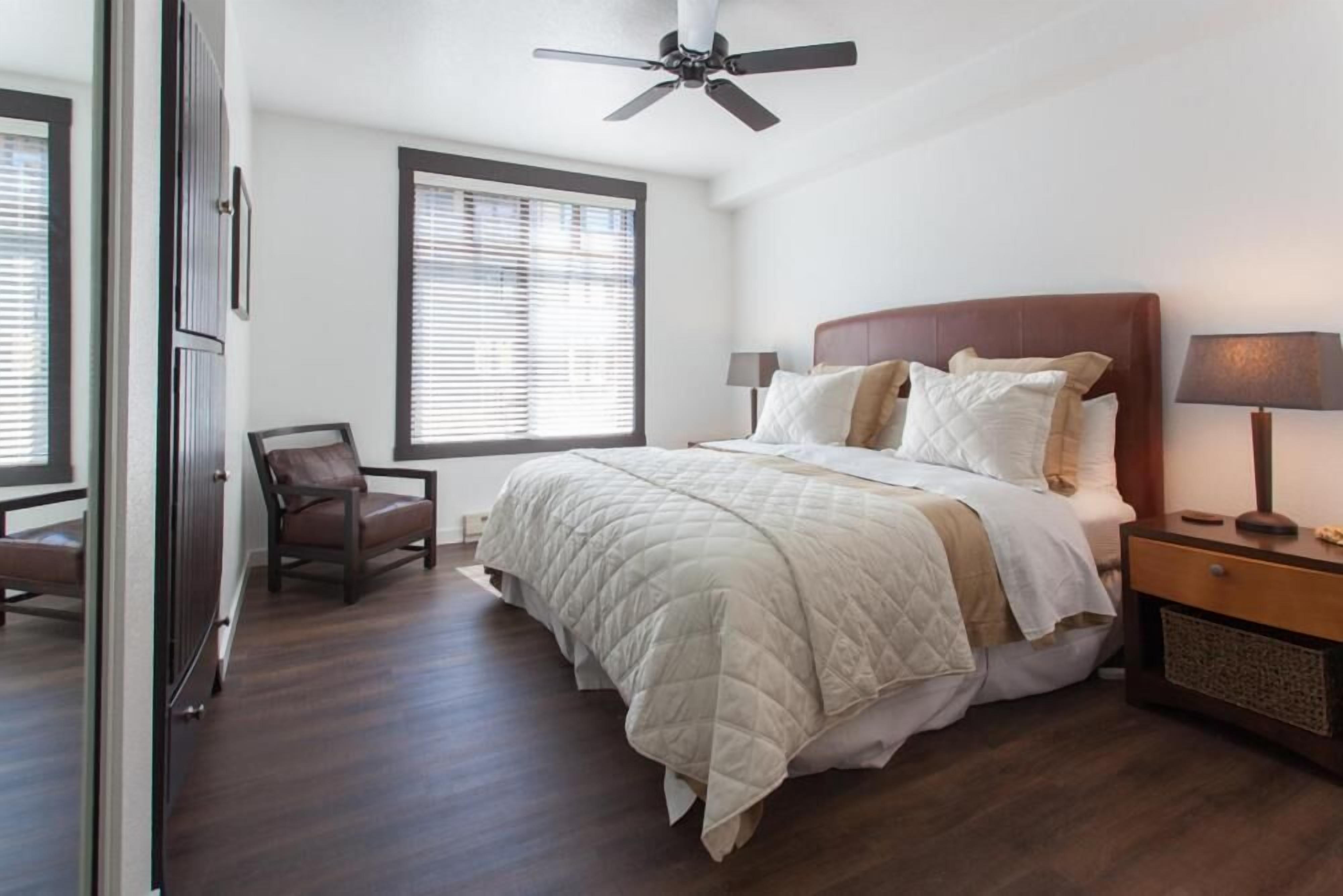 A bedroom with a bed, a chair, a nightstand, a ceiling fan, and a window with blinds.