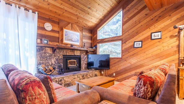 A cozy cabin interior with a stone fireplace, wooden walls, a large window, and a television. - Snowflower 62 (Mammoth Lakes)