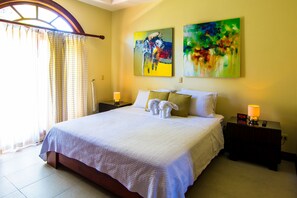 A neatly made bed with white linens, two pillows, and a set of towels. There are two bedside tables with lamps and a clock. The room has a large window with curtains, and two abstract paintings on the wall.