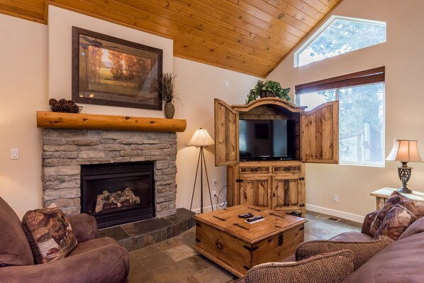 A cozy living room with a stone fireplace, wooden entertainment center, and comfortable seating. - Mammoth Gateway Village 11 (Mammoth Lakes)