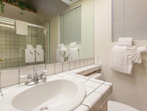 Ein Badezimmer mit einem großen Spiegel, einem Waschbecken und einer Toilette. Mehrere Handtücher hängen an der Wand und ein Fenster mit Jalousien.