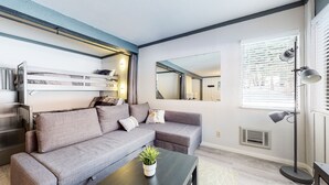 Un saló modern amb un sofà gris, una taula baixa amb una planta, un llit superposat i un mirall gran que reflecteix l'habitació.