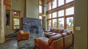 A spacious living room with a stone fireplace, leather sofas, and large windows offering a view of the outdoors.