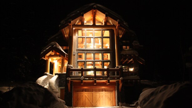 A wooden cabin with a large central window and a garage door, surrounded by snow.