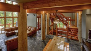 A spacious living area with a stone floor, wooden beams, a staircase with a wrought iron railing, and a large window offering a view of the outdoors.