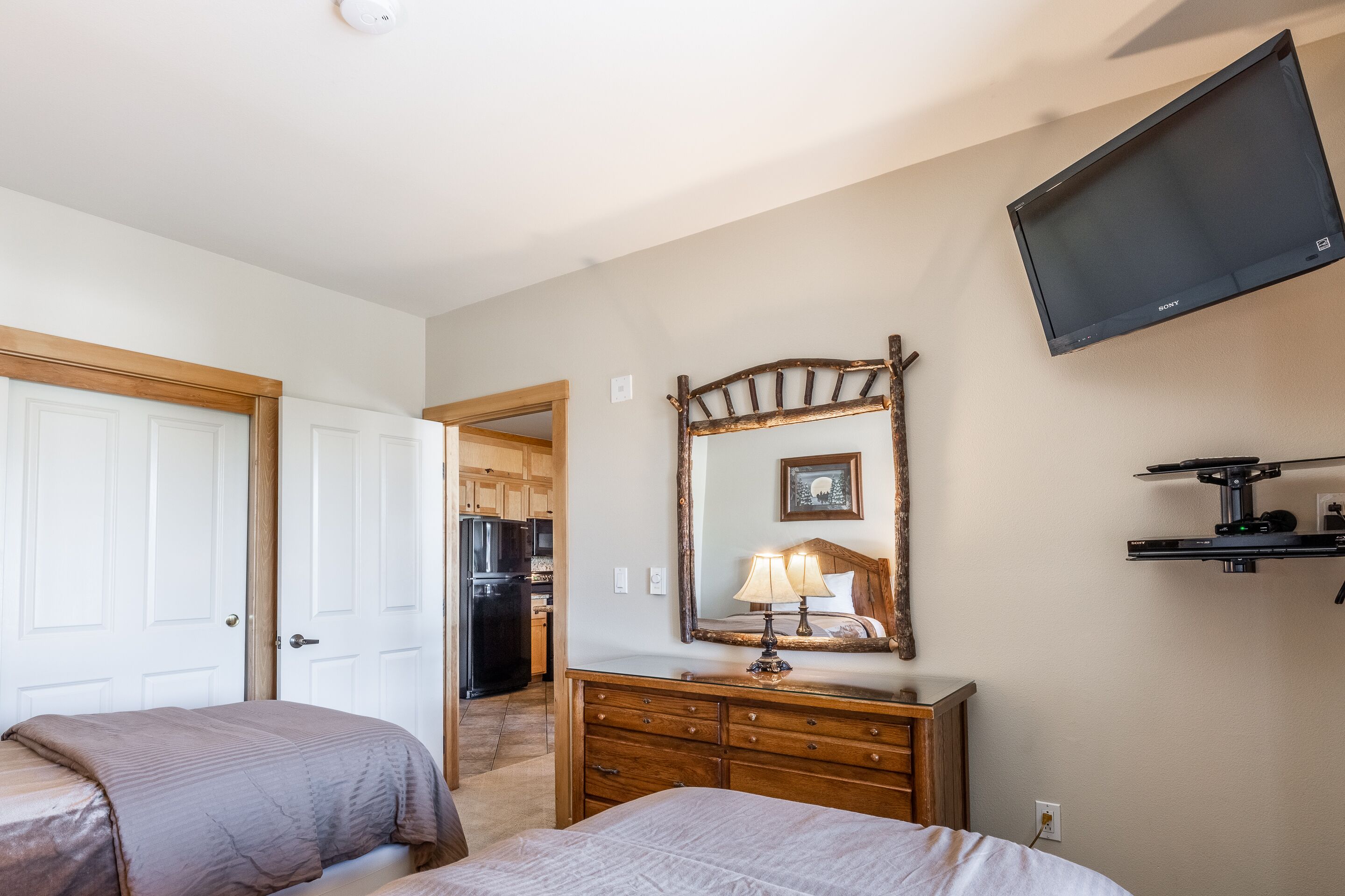 A hotel room with two beds, a dresser, a mirror, a television mounted on the wall, and a kitchen area visible through an open door.