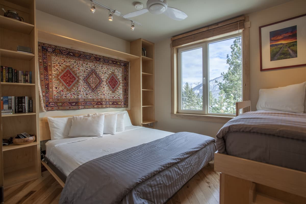 Un dormitorio con una cama grande, una estantería, una ventana con vistas a árboles y un ventilador de techo.