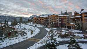 Eine mehrstöckige Lodge vor einer verschneiten Landschaft, einer gewundenen Straße und einem Bergpanorama.