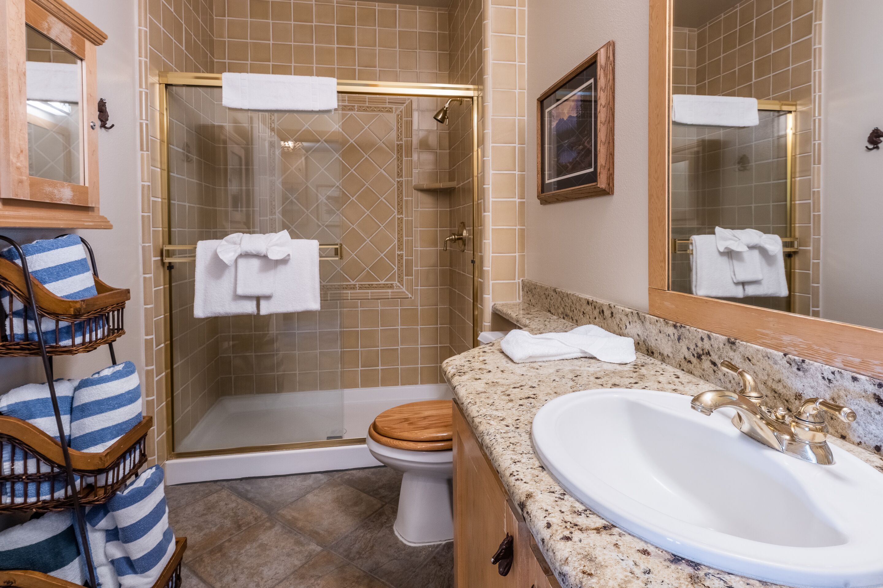 A bathroom with a glass-enclosed shower, a wooden toilet, a sink with a gold faucet, and a towel rack with striped towels.