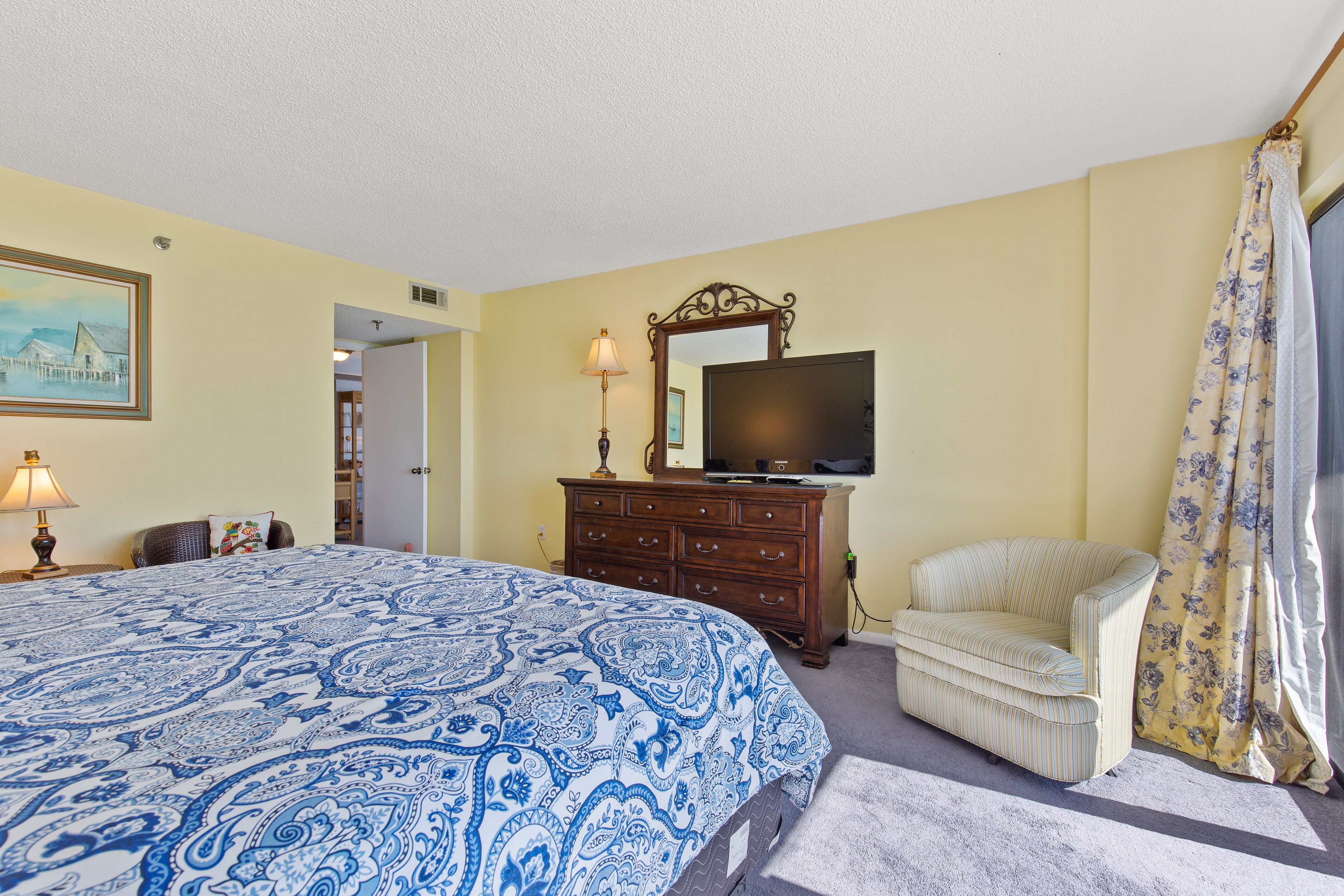 Une chambre à coucher avec un grand lit, une commode, une chaise, un téléviseur et un tableau accroché au mur.