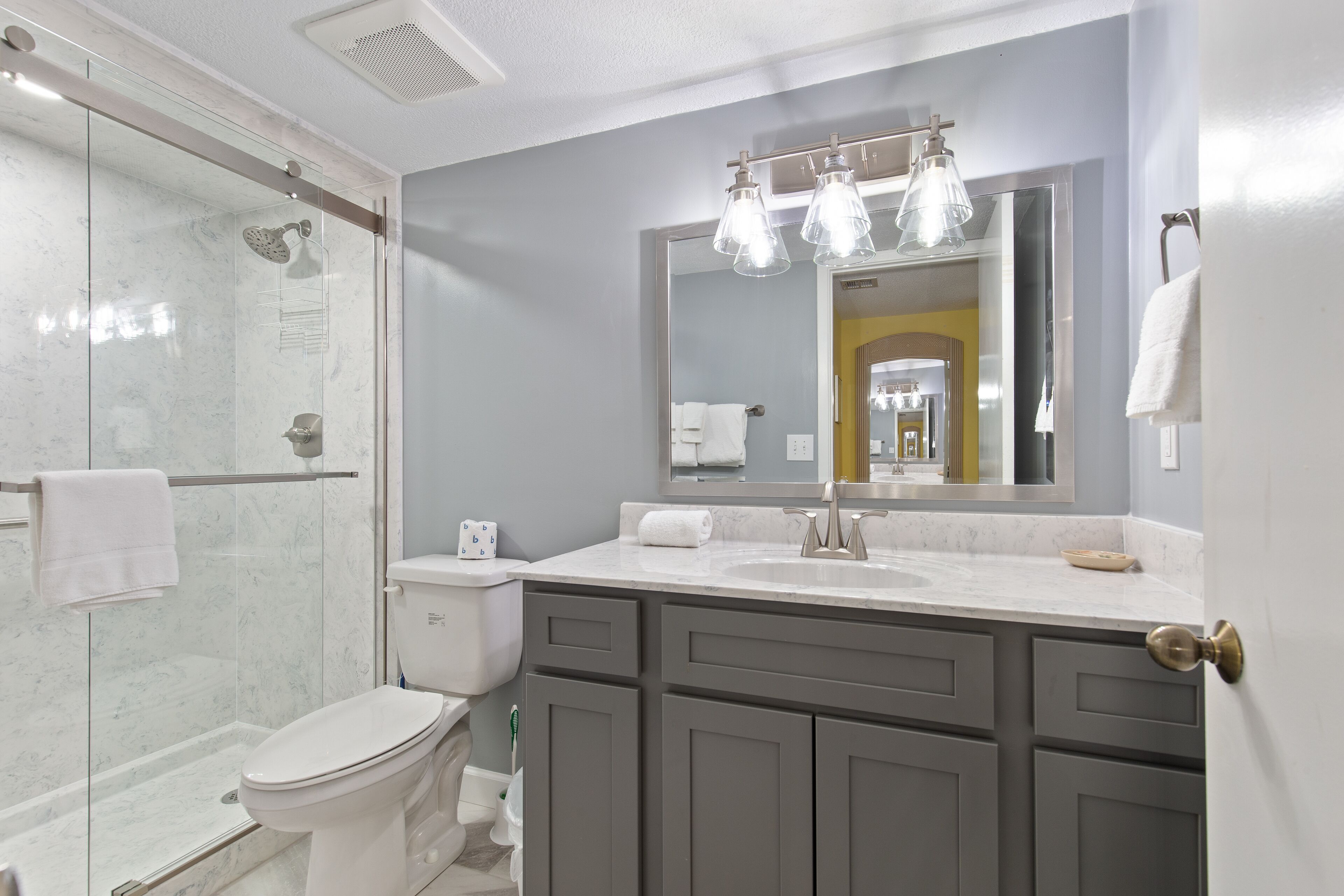 A modern bathroom with a glass-enclosed shower, a white toilet, and a vanity with a large mirror and overhead lighting.