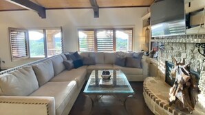 A living room with a sectional sofa, a glass coffee table, a stone fireplace, and large windows with blinds. - The LonePine in YellowHouse Canyon (Slaton)