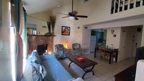 Un salon avec un canapé, des fauteuils, une table et une cheminée. On peut apercevoir un ventilateur de plafond, un tableau et une cuisine à l’arrière-plan.