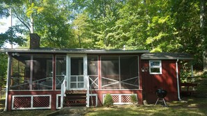 Une cabane rouge avec une véranda grillagée, une petite terrasse et un barbecue, située dans une zone boisée.