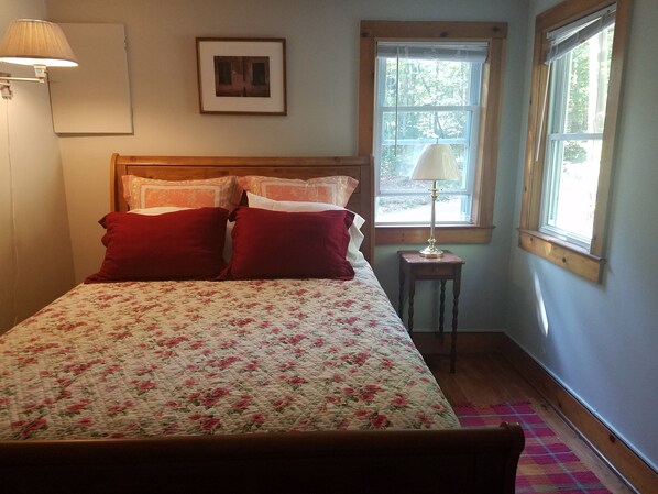 A bedroom with a floral bedspread, a wooden headboard, two red pillows, a bedside table with a lamp, and a window with a view of trees.