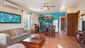 A living room with a sofa, coffee table, and dining area with chairs. There is a kitchen area with cabinets and a refrigerator.