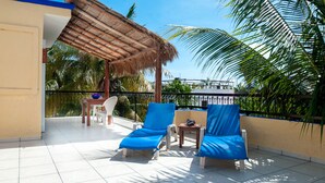A balcony with two blue lounge chairs, a small table with a bowl, and a thatched roof structure.