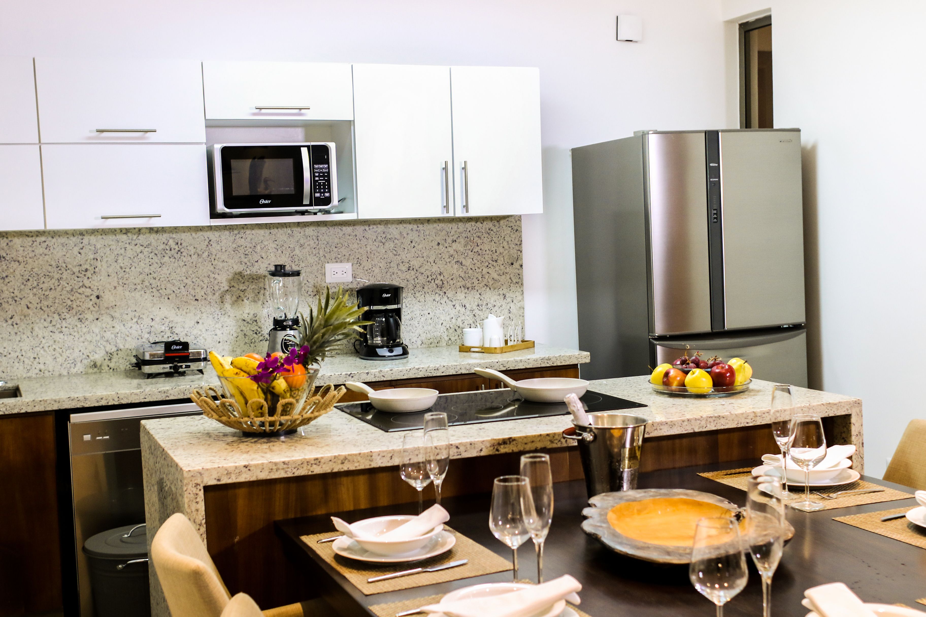 Una cocina moderna con armarios blancos, microondas, refrigerador de acero inoxidable y un comedor dispuesto con platos, vasos y frutero.