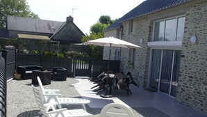 Un espace extérieur aménagé en terrasse, avec des chaises et des tables en plastique blanc, un bâtiment en pierre doté d’un toit en tuiles et une clôture noire.