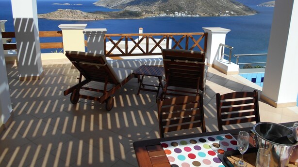 Um terraço com duas cadeiras de madeira e uma mesa, com vista para a costa, uma montanha imponente e o mar.