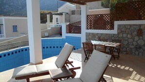 Área da piscina com espreguiçadeiras e uma mesa, uma parede de pedra e vista para edifícios ao fundo.