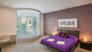 A modern bedroom with a large bed, a built-in bathtub, and a view of greenery.