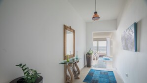 A hallway with a mirror, a glass console table, a potted plant, a blue rug, and a painting on the wall.