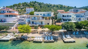A coastal hotel with multiple buildings, a clear pool area with sun loungers, and a hillside backdrop. - Villa La Perla - beachfront villa for ten, private pool and amazing views by MyWaycation (Rogoznica)