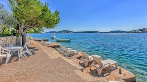 A pebble beach with lounge chairs and a stone wall, overlooking a calm sea with boats. - Villa La Perla - beachfront villa for ten, private pool and amazing views by MyWaycation (Rogoznica)