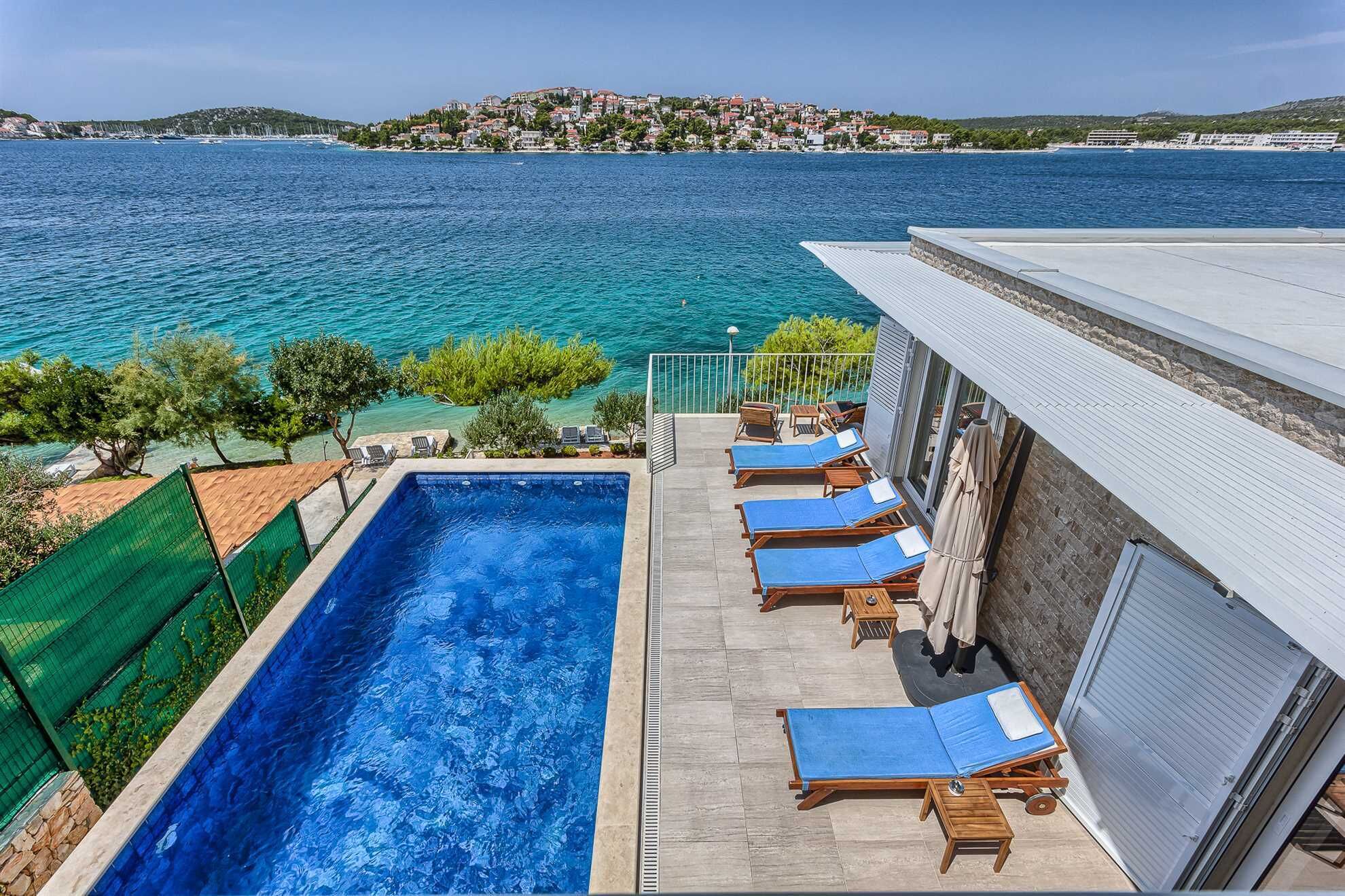 A pool area with lounge chairs and a view of the sea and a distant town.