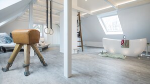 A modern, spacious room with a sloped ceiling, a large skylight, a wooden stool, a white bathtub, and a bench with cushions. - Design - Studio in Gütersloh with balcony (Gütersloh)