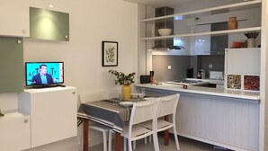 A modern kitchen with white cabinetry, a dining area with a table set for two, and a television displaying a show. - <--Right on this BEACH, Bright 1-Bedroom Apartment; OCEAN VIEW. ****42 m² + TERRACE + garage + Wi-Fi (Capbreton)