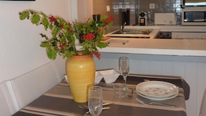 A dining area with a table set for two, a vase with flowers, and a kitchen in the background. - <--Right on this BEACH, Bright 1-Bedroom Apartment; OCEAN VIEW. ****42 m² + TERRACE + garage + Wi-Fi (Capbreton)