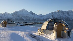 Deux structures en forme de dôme, recouvertes de neige et dotées de toits transparents, se dressent sur un sol enneigé, avec en arrière-plan des montagnes aux sommets enneigés et une étendue d’eau.