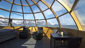 Une pièce circulaire et chaleureuse, dotée d’un plafond en verre qui offre une vue panoramique sur des montagnes enneigées et un plan d’eau.