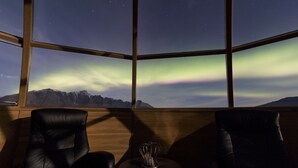 Une pièce dotée d’une grande fenêtre offrant une vue sur les aurores boréales qui illuminent un paysage montagneux.