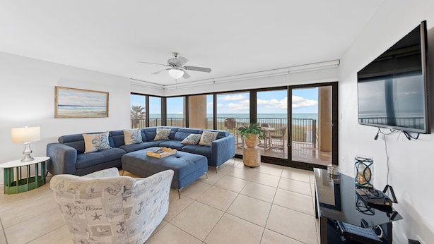 A spacious living room with a large sectional sofa, a small round table, a flat-screen TV mounted on the wall, and a balcony with a view of the ocean. - Walls of windows overlooking the blue ocean!  (South Padre Island)