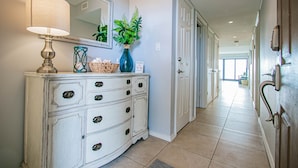 A hallway with a white dresser, a mirror, a lamp, and a vase with green plants. - Walls of windows overlooking the blue ocean!  (South Padre Island)