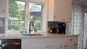 A kitchen with a large window, a sink, a dishwasher, and a countertop with a bottle of wine. - Falmouth - walk to beach, walking path, close to downtown, private yard (Falmouth)