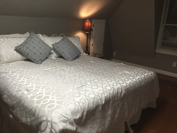 Ein Schlafzimmer mit einem Muster-Bettbezug, zwei Kissen, einer Nachttischlampe und einem Fenster mit Jalousien.