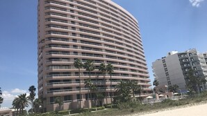 A tall, curved hotel building with multiple balconies, located near a beach with palm trees and other buildings in the background.