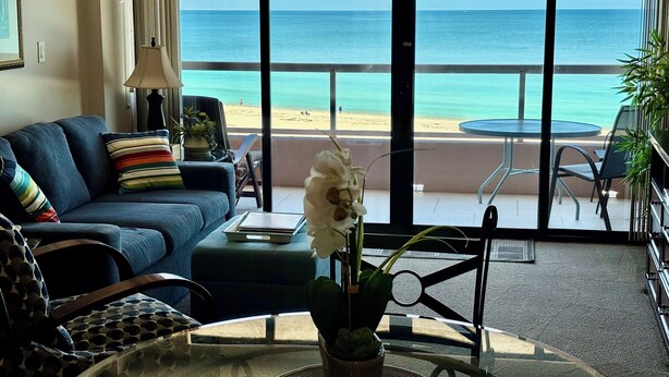 A living room with a sofa, a table with a vase, and a view of the beach and ocean through large windows.