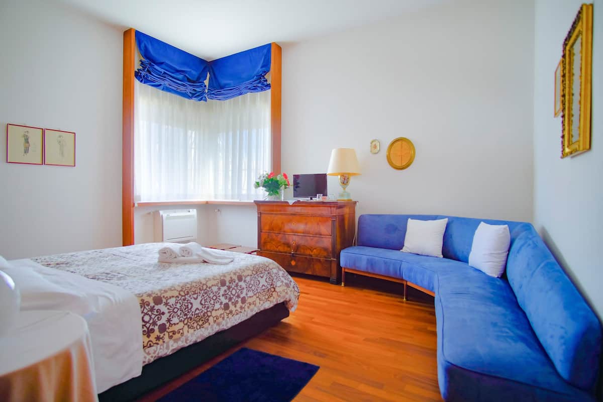 Een slaapkamer met een bed, een blauwe bank, een houten dressoir en een raam met gordijnen.