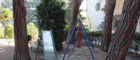 A playground with a slide and swings, surrounded by trees and a building in the background.