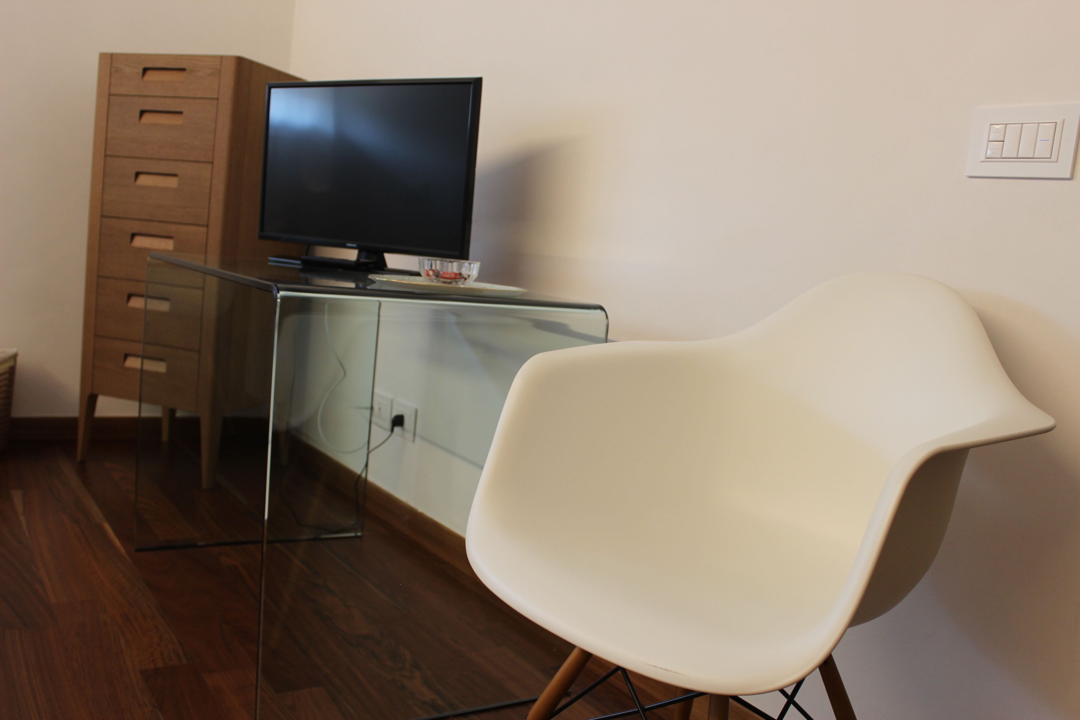 Une chambre moderne avec un bureau en verre, un téléviseur à écran plat, une chaise blanche et un meuble en bois.