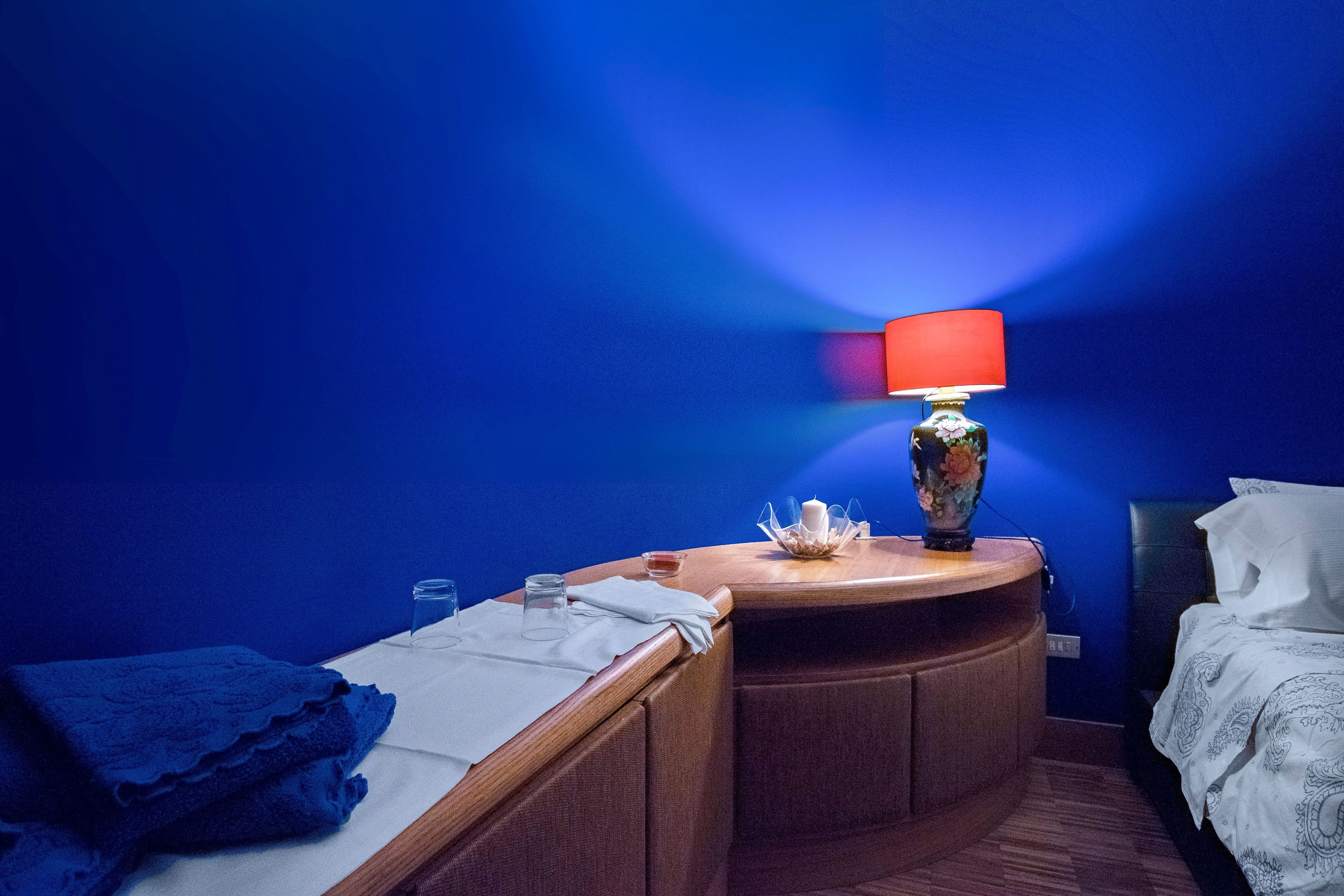 Quarto de hotel com secretária de madeira curva, luminária vermelha, um vaso e cama com roupa de cama branca.