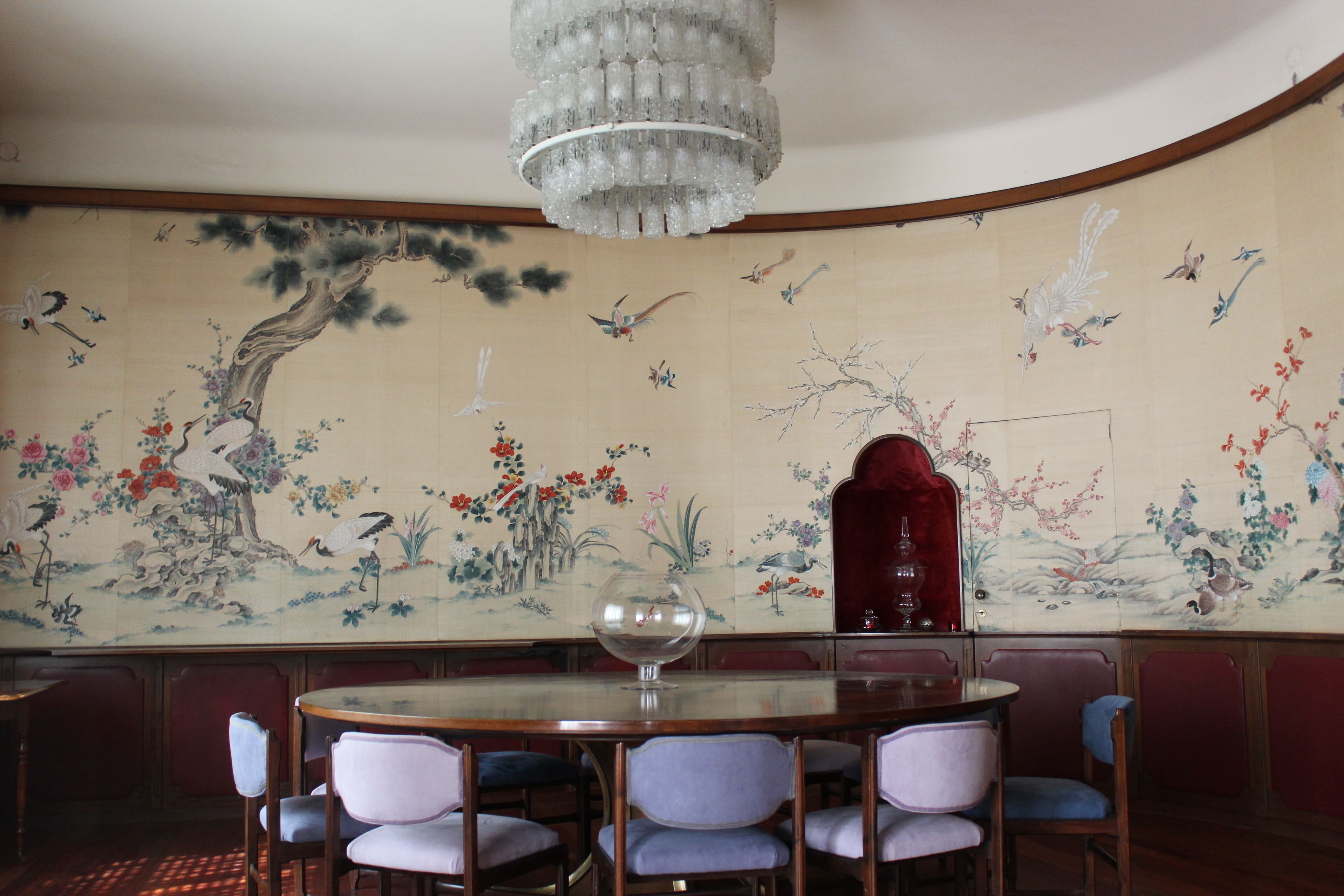 A traditional room with a round table and chairs, a chandelier, and a wall with a detailed mural.
