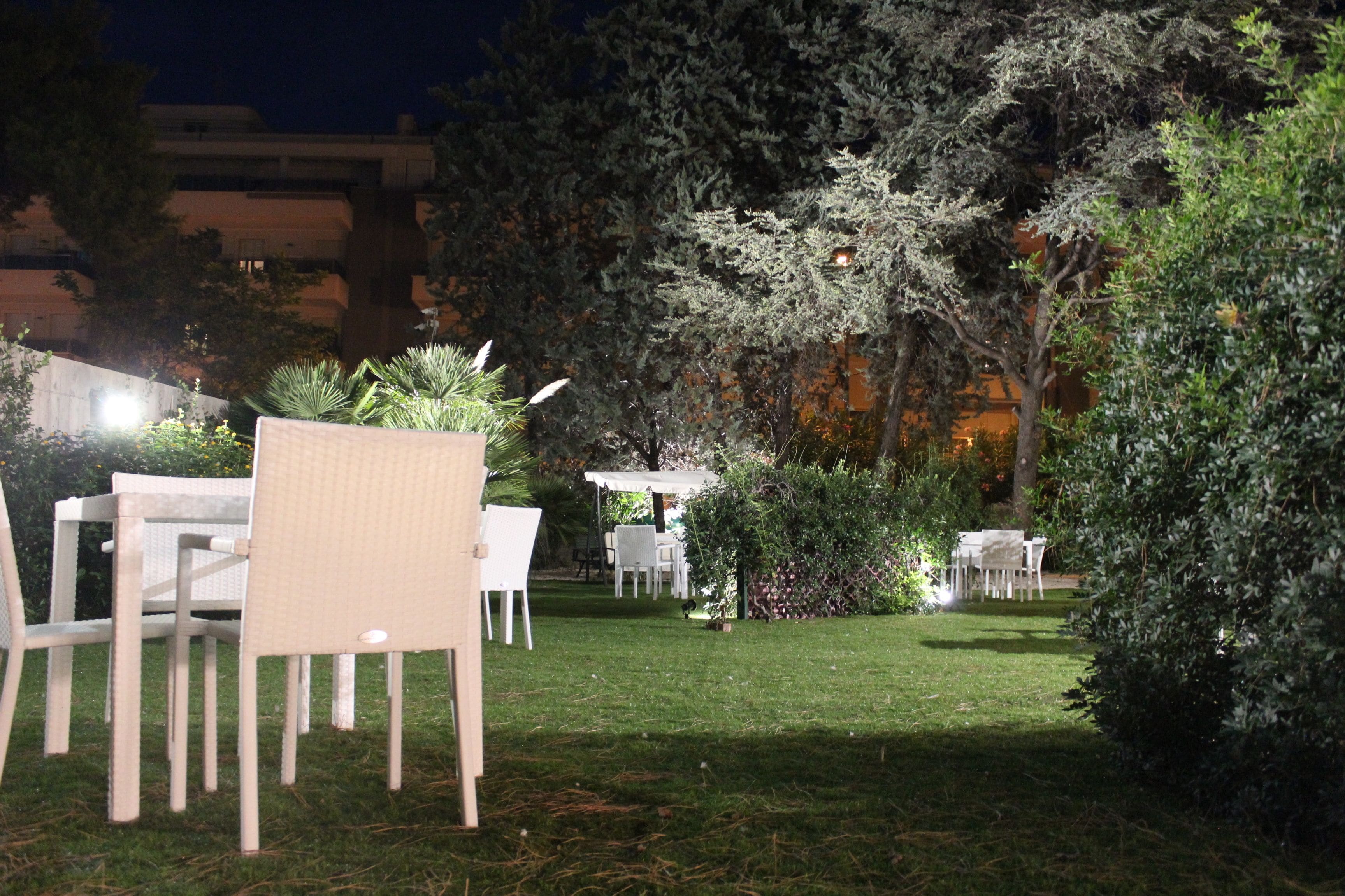 Un espace de restauration en plein air avec des tables et des chaises blanches, entouré de verdure et d’arbres.
