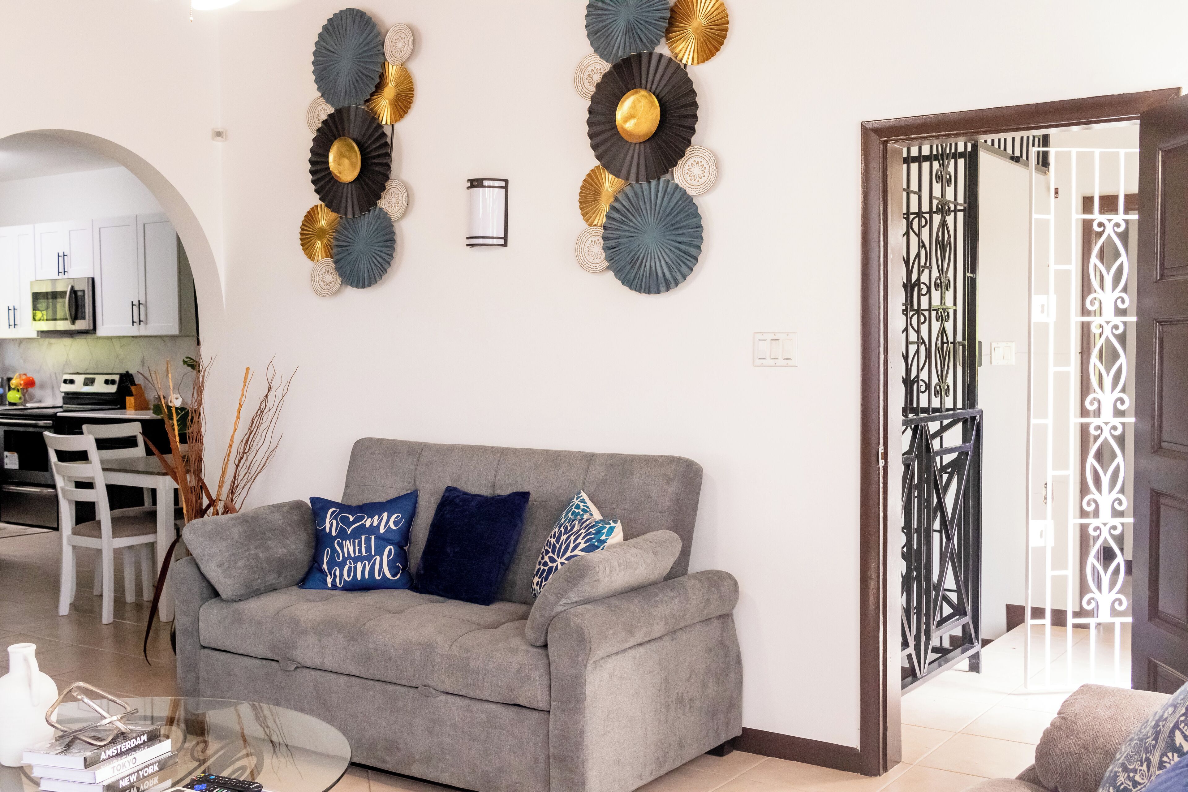 Una sala de estar con un sofá gris, decoración en la pared, una mesa de centro de vidrio y vista a la cocina al fondo.