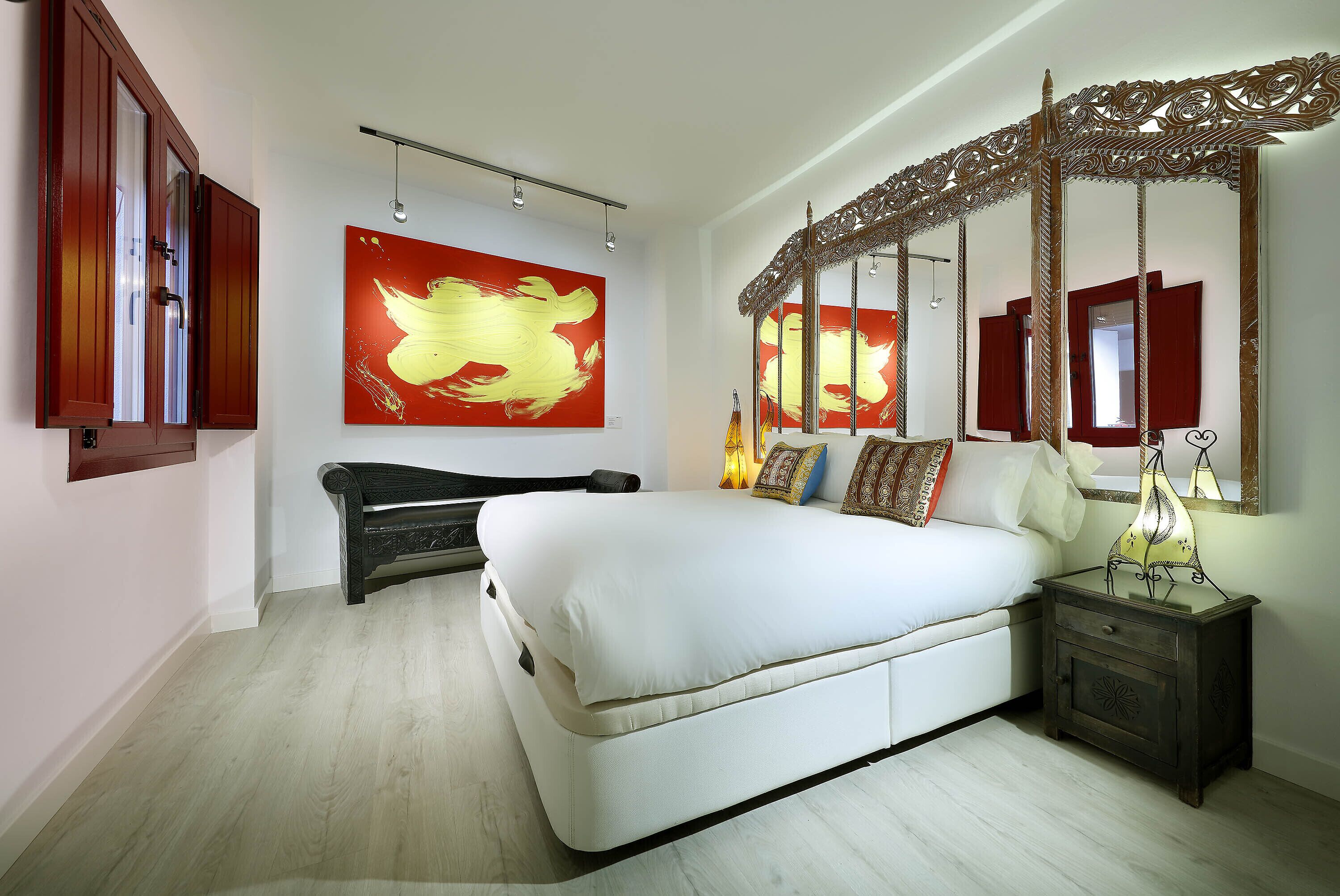 Een slaapkamer met een groot bed, een nachtkastje, een spiegel en een abstract schilderij in rode tinten aan de muur.